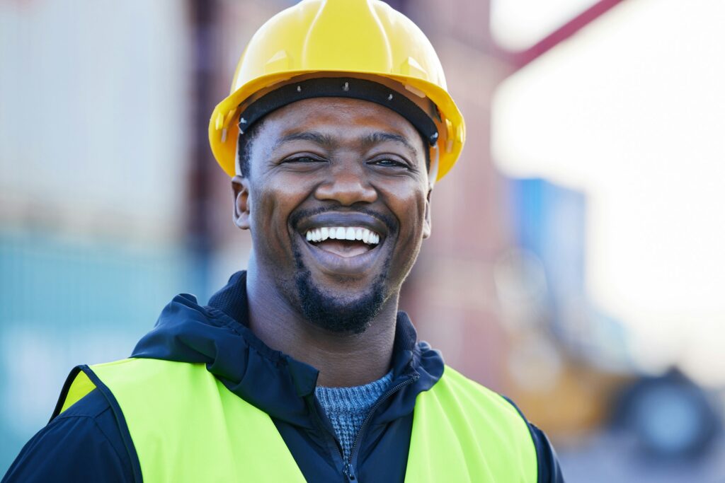 Portrait, logistics and black man smile with helmet or hard hat at shipyard and confident at work.